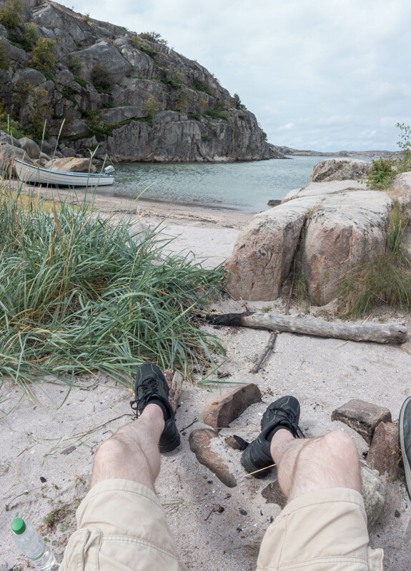 Vy över en sandstrand på ön Trinisla på västkusten. Man ser bloggägarens ben klart och tydligt, där fötterna vilar på en grillplats.