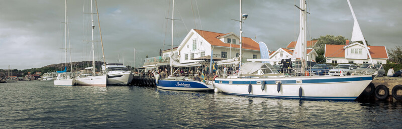 Vy över restaurangen Hjalmars på Hamburgsund på västkusten. Flera båtar ligger dockade utanför restarurangen och många sitter och äter i kvällssolen.