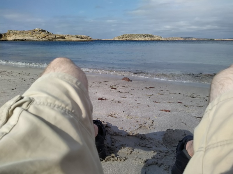 Vy över en strand på Vedholmen på västkusten med bloggägarens ben synliga, sittandes med shorts, strumpor och skor.